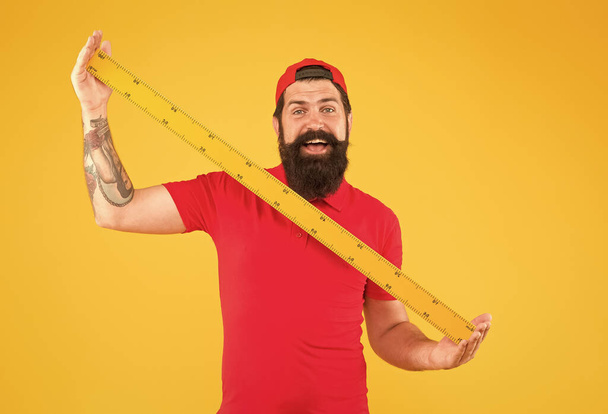 Post office worker with ruler. Big size. Measuring tool. Man holding ruler on yellow background. Bearded man preparing for maths. Hipster man in cap with big measuring instrument. Size of parcel - Foto, immagini