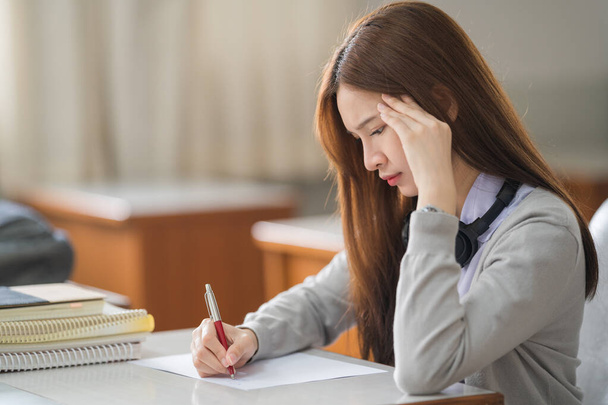 Çaresiz ve stresli Asyalı kız öğrenci üniforması içinde üniversite sınıfında tek başına sınav ödevi yapıyor. - Fotoğraf, Görsel