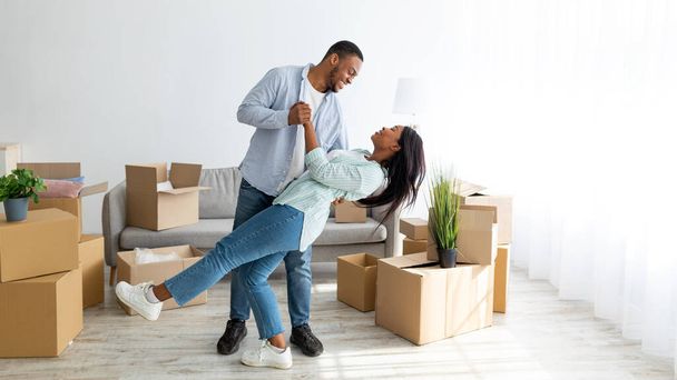 Feliz pareja afroamericana cariñosa bailando en su nueva casa entre cajas de cartón en día de mudanza, panorama - Foto, Imagen