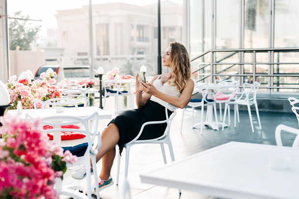 Een aantrekkelijke vrouw zit in een café en gebruikt haar telefoon om berichten uit te wisselen en kijkt bedachtzaam in de verte. Moderne technologieën, online communicatie - Foto, afbeelding