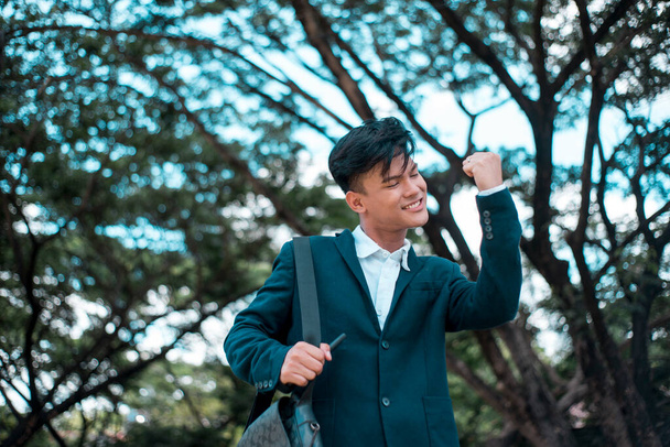 A happy young asian man with hands raised in a gesture of success. Passing an exam, getting good grades at school, got the girl of his dreams, or getting accepting at work. Good news concept. - Photo, Image