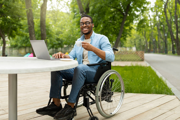 Etätyö vammaisille. Iloinen musta kaveri pyörätuolissa työskentelee verkossa, käyttäen kannettavaa ulkona kahvilassa - Valokuva, kuva