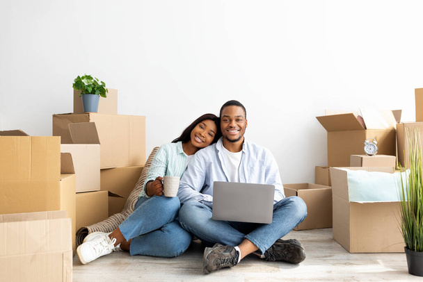 Conceito comovente. Feliz casal afro-americano comprando móveis para novo apartamento, sentado no chão e usando laptop - Foto, Imagem