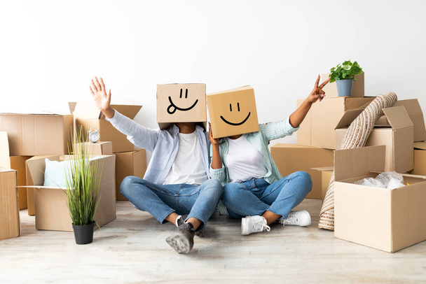 Концепция переезда. Игривая пара с картонными коробками с улыбающимися лицами на голове, сидя на полу и веселясь - Фото, изображение