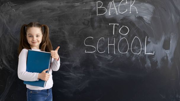 Молодая кавказская школьница стоит на фоне меловой доски. Слова написаны обратно в школу. держит в руках несколько больших блокнотов и большой палец вверх. - Фото, изображение