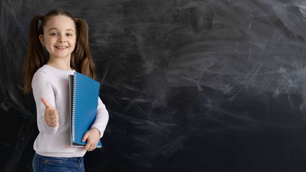a Caucasian girl, a schoolgirl, is standing against the background of an empty chalk board. Holds a notebook in his hands. The thumb raises up. - Photo, image
