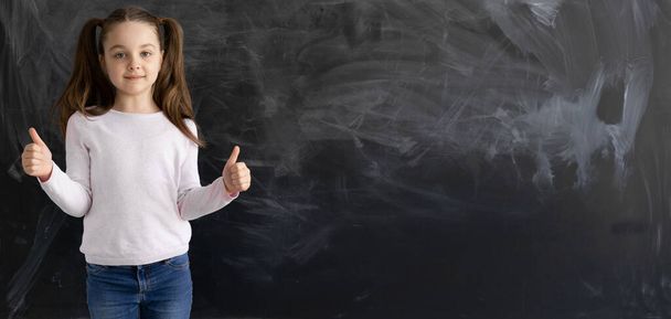 a Caucasian girl, a schoolgirl, is standing against the background of an empty chalk board. The thumb is raised up. - Фото, изображение