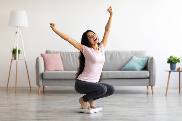 Excited Indian woman sitting on scales, raising hands up, happy with result of her weight loss program at home - Foto, Imagen