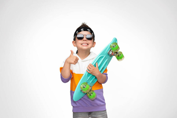 Kleiner cooler Skaterboy mit stylischer Kleidung und Sonnenbrille hält einen türkisfarbenen Schlittschuh in der Hand und zeigt den Daumen nach oben. - Foto, Bild