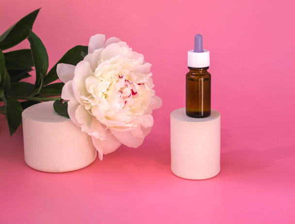 Composition of dark glass with cosmetic, medical products. Bottles with allergy medicine or serum, oils on catwalks with white peony on pink background. - Photo, Image