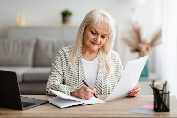 Smiling mature woman writing and holding document at home - Photo, Image