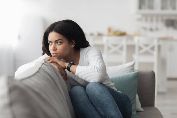Mulher afro-americana solitária deprimida sentada no sofá com expressão facial triste e olhando para o lado, espaço livre - Foto, Imagem