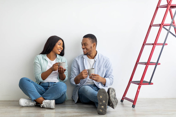 Young african american couple discussing plan of repairs in their new house and drinking coffee, sitting on floor - Fotó, kép