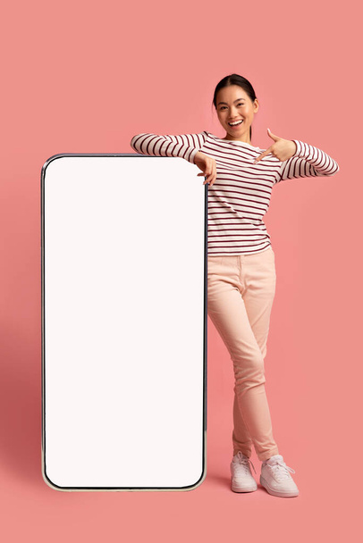 Alegre señora asiática inclinándose y señalando Smartphone grande con pantalla blanca - Foto, imagen