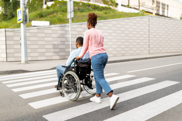 Widok z tyłu młoda opiekunka pchanie wózka inwalidzkiego z czarnym pacjentem płci męskiej całej ulicy miasta, pełna długość - Zdjęcie, obraz