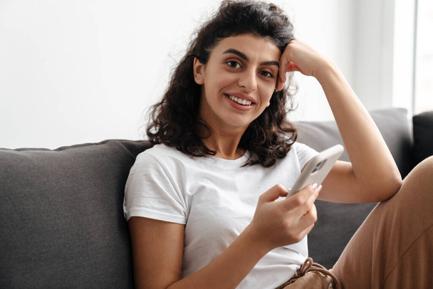 gelukkig brunette jong vrouw met behulp van mobiele telefoon terwijl zitten op de bank thuis - Foto, afbeelding