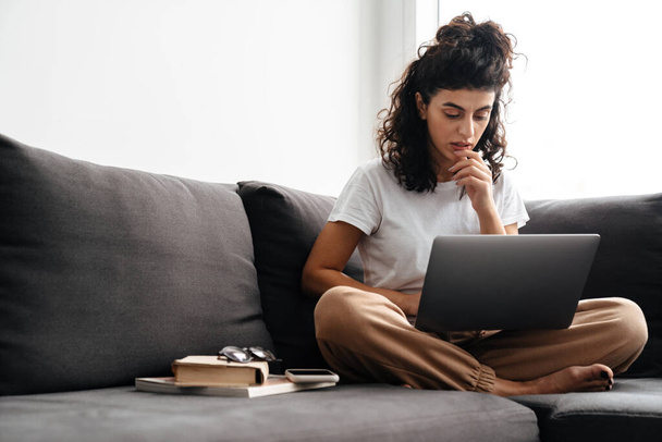 Focused young woman working with laptop while sitting on sofa at home - Φωτογραφία, εικόνα