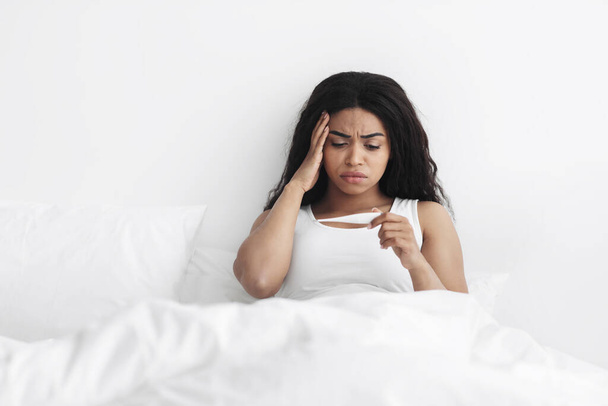 Hohes Fieber, Unwohlsein. Kranke schwarze Frau hält Thermometer in der Hand, überprüft Körpertemperatur im Bett - Foto, Bild