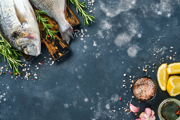 Raw dorado fresh fish or sea bream with ingredients for making lemon, thyme, garlic, cherry tomato and salt on a black slate, stone or concrete background. Top view with copy space. - Фото, изображение