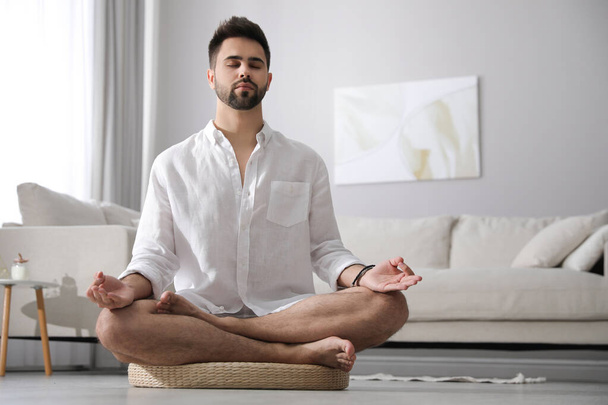Young man meditating on straw cushion at home, space for text - Photo, image