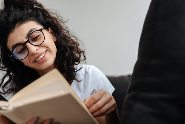 Glimlachende jonge vrouw in bril leesboek terwijl ze thuis op de bank rust - Foto, afbeelding