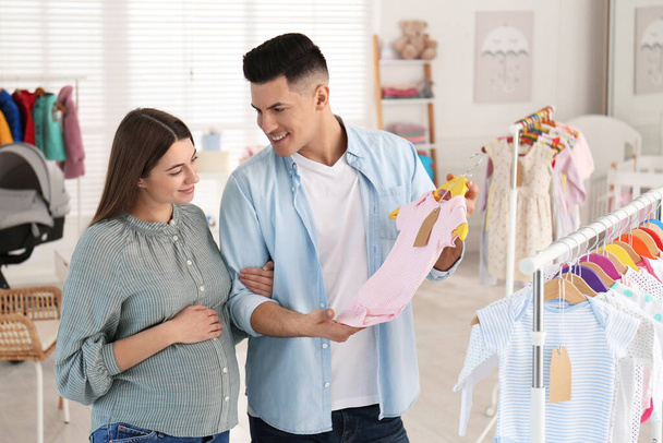 Happy pregnant woman with her husband choosing baby clothes in store. Shopping concept - Foto, Imagem