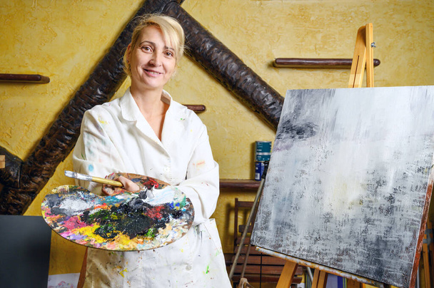 Ικανοποιημένη γυναίκα καλλιτέχνης ποζάρει κοντά σε νέα εικόνα δίπλα στο easel. - Φωτογραφία, εικόνα