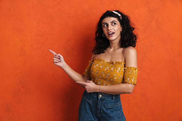 Attractive smiling young woman pointing finger at copy space isolated over orange background - Photo, image