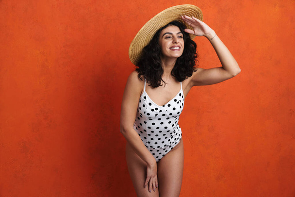Joyful beautiful woman in swimsuit smiling while looking aside isolated over orange background - 写真・画像