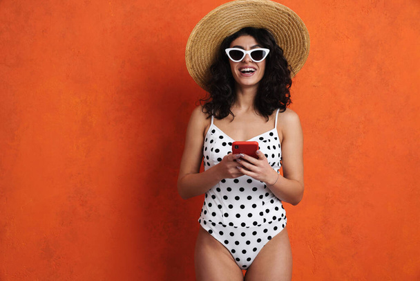 Joyful beautiful woman in swimsuit smiling while using cellphone isolated over orange background - Photo, Image