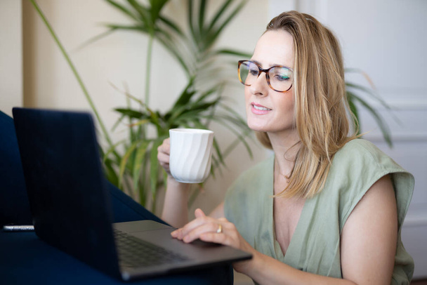 Aantrekkelijke jonge vrouw werkt op laptop en het drinken van koffie terwijl zitten op de vloer thuis - Foto, afbeelding