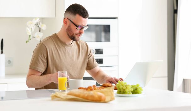 Jovem focado homem bonito vestindo óculos trabalhando remotamente no laptop enquanto sentado à mesa na cozinha moderna em casa, freelancer masculino saudável tomando café da manhã e olhando para o computador - Foto, Imagem