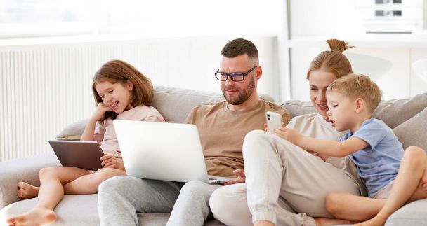 Fiatal, boldog család, szülők két gyerekkel, akik modern technológiákat használnak, miközben együtt töltik az időt otthon. Apa, anya és gyerekek laptop, digitális tabletta és okostelefon pihentető kanapén - Fotó, kép