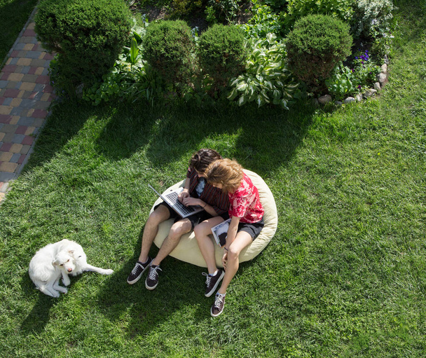 teenagers 16-17 years old, a guy and a girl, are sitting in a large soft pouf on grass, looking at screen on laptop. online communication, distance learning, exam preparation, modern technologies - Photo, image