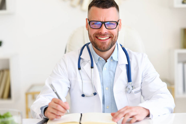 Médico masculino amigável alegre vestindo casaco branco com estetoscópio nos ombros olhando para a câmera com sorriso enquanto trabalhava no hospital, médico masculino sentado em seu local de trabalho e esperando pelo paciente - Foto, Imagem