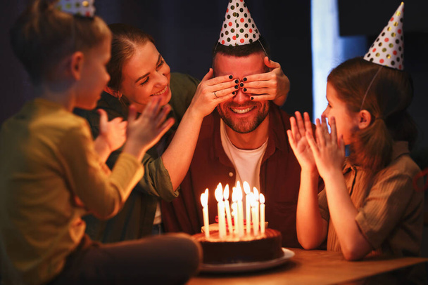 Time to make a wish. Young happy beautiful family with children wearing party hats congratulating father with Birthday at home. Excited dad with covered eyes going to blow candles on chocolate cake - Photo, Image