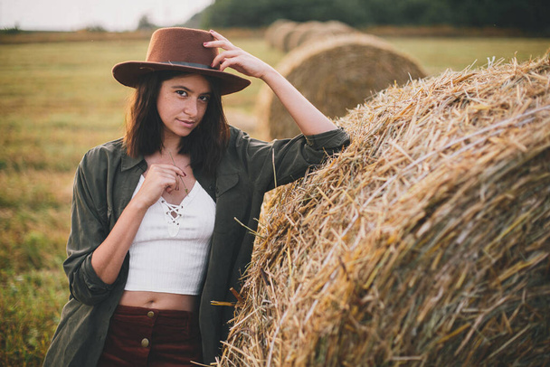 藁と帽子の美しいスタイリッシュな女性は夏の夜のフィールドで干し草をポーズ。セクシーな若い女性リラックスで干し草のベール,田舎の夏休み. - 写真・画像