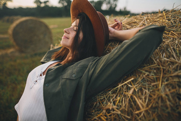 夏の夜のフィールドで干し草の上でリラックス帽子の美しいスタイリッシュな女性。干し草の玉で夕日を楽しむ魅力的な若い女性の肖像画。田舎の静かな瞬間 - 写真・画像