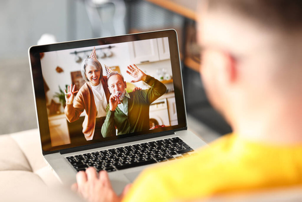 Pozytywna para starszych rodziców w kapeluszach imprezowych gratuluje dorosłemu synowi online przez wideo połączenie na laptopie, macha przed kamerą i gwiżdże, człowiek mający wideo połączenie podczas świętowania urodzin w domu - Zdjęcie, obraz