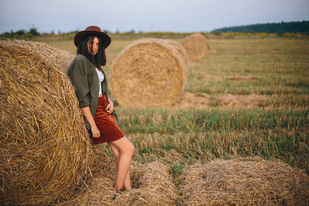 夏の夕方に畑の干し草の俵に立つ帽子のスタイリッシュな女性。落ち着いた雰囲気の瞬間。若いファッショナブルな女性は、田舎の夏休み、干し草の山でリラックスします。農村のスローライフ - 写真・画像