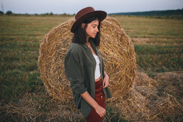夏の夕方に畑の干し草の俵に立つ帽子のスタイリッシュな女性。落ち着いた雰囲気の瞬間。若いファッショナブルな女性リラックスでhaystack,田舎の休暇 - 写真・画像