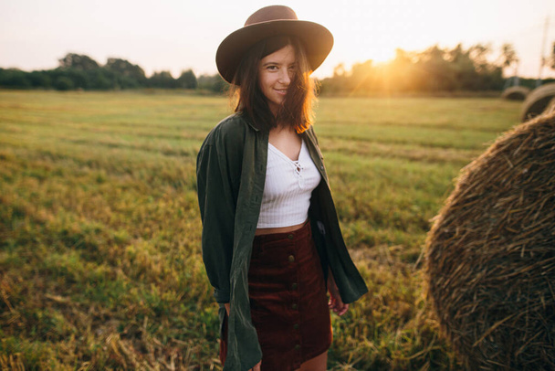 Beautiful stylish woman in hat walking at haystacks in sunset light in summer field. Atmospheric tranquil moment in countryside. Young female enjoying evening at hay bale in warm sunshine - 写真・画像