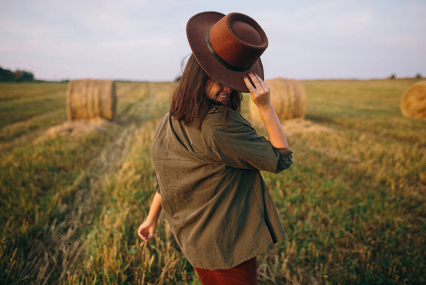 Şapkalı güzel bir kadın yaz tarlasında yürüyor ve gülümsüyor. Genç ve mutlu bir bayan kırsalda dinleniyor, akşamın tadını çıkarıyor. Atmosferik an. - Fotoğraf, Görsel