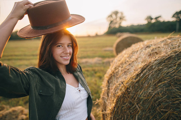 美しい屈託のない女性は、夕方の夏のフィールドの干し草で日没の光の中で帽子を引く。田舎の干し草の山でリラックスしている若い幸せな女性。大気の瞬間 - 写真・画像