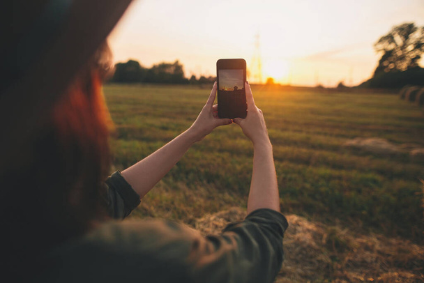 夏のフィールドで携帯電話で夕日の写真を撮るスタイリッシュな女性。若い女性はスマートフォンを持ち、田舎で夜の暖かい日差しを捉えます。大気中の美しい瞬間 - 写真・画像