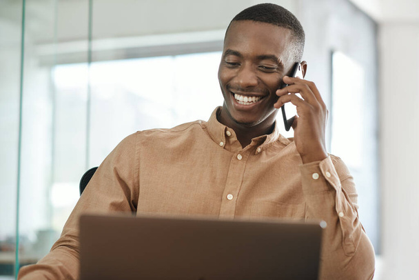 若いアフリカの実業家が携帯電話で話をし、彼のオフィスの机に座っている間にノートパソコンで作業 - 写真・画像