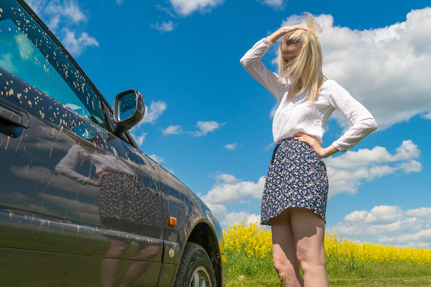 slender young blonde woman in a skirt and white blouse stands near a dirty car stuck on a dirt road in the middle of a rapeseed field, looks at the wheel and holds her head - Foto, Bild