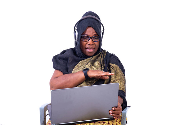 hermosa mujer de negocios musulmana sentada sobre fondo blanco comunicándose con el uso de ordenador portátil y auriculares con mirada enojada. - Foto, imagen