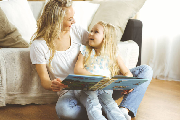 Happy family. Blonde young mother reading a book to her cute daughter while sitting at wooden floor. Motherhood concept - Fotoğraf, Görsel
