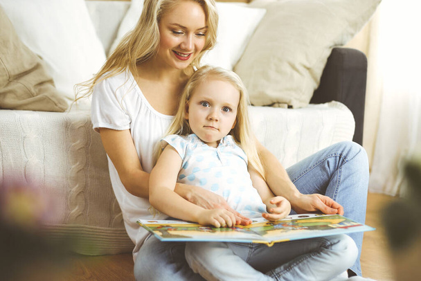 Una famiglia felice. Giovane madre bionda che legge un libro alla sua graziosa figlia mentre è seduta sul pavimento in legno. Concetto di maternità - Foto, immagini
